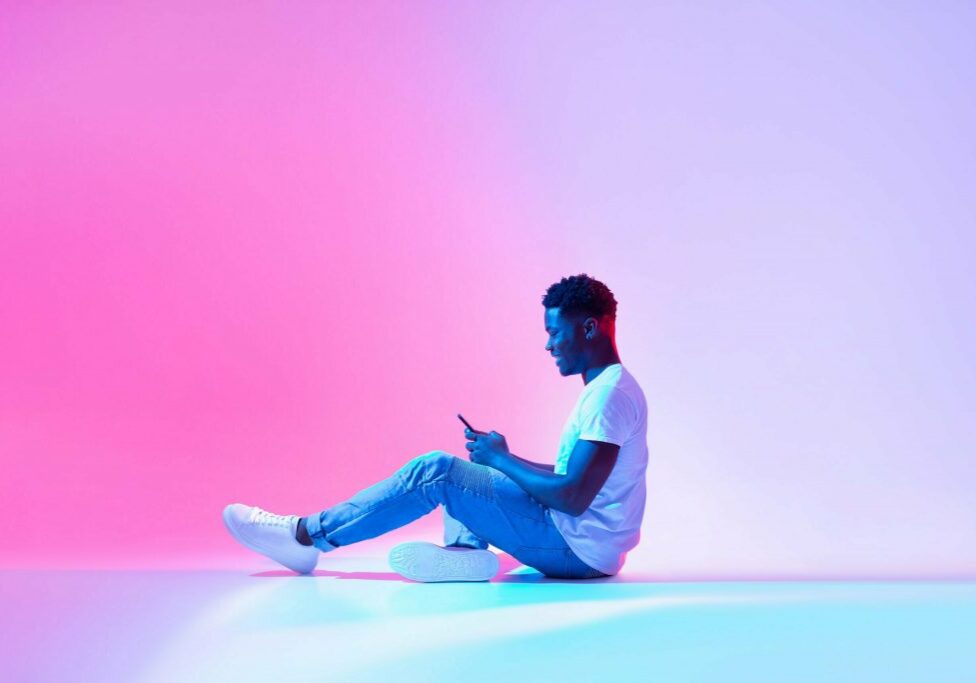 Cool mobile app. Side view of happy black man using modern smartphone in neon light, copy space. Positive African American guy working or studying online, browsing social network, chatting on web