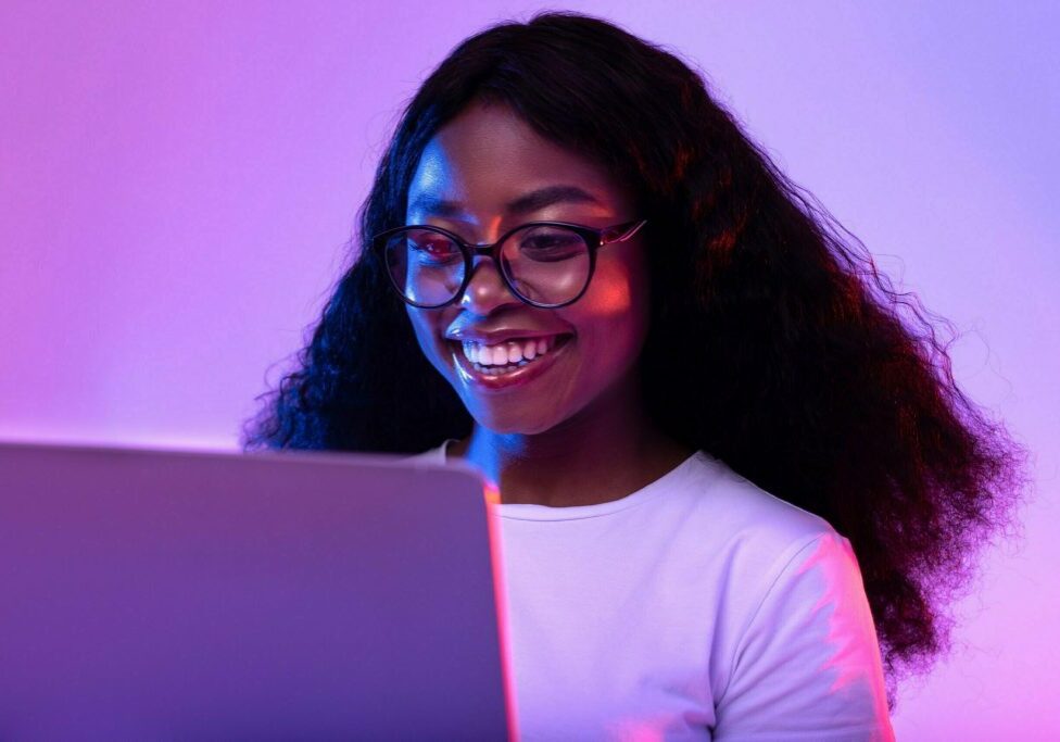 Happy young black woman in glasses using laptop pc for online work or studies in neon light. Cheery African American lady having remote video conference, chatting with her friends on web